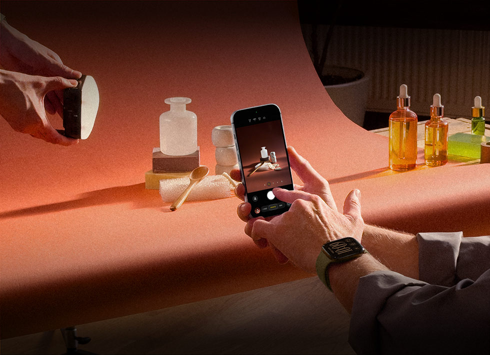 A photoshoot in a workplace setting, one person is using an iPhone to photograph products against a backdrop, one person is holding a light